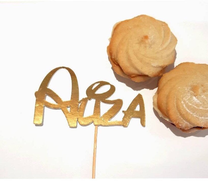 Puede incluir: Topper de pastel de purpurina dorada con el nombre "Alba" en un palo de madera. Dos galletas de mantequilla con glaseado blanco est&aacute;n en el fondo.