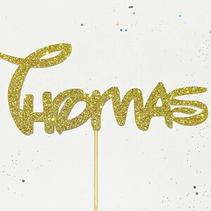 Puede incluir: Topper para pastel de purpurina dorada con el nombre "Thomas" en una fuente caprichosa.