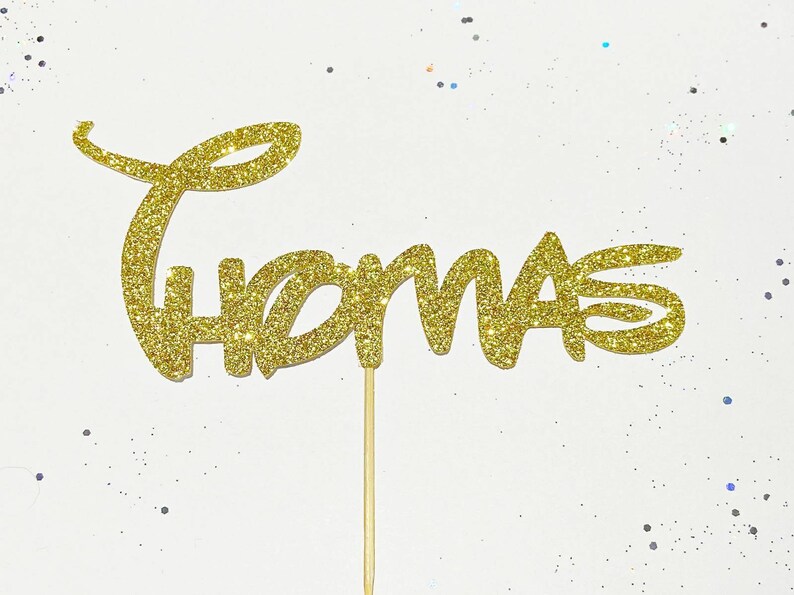 Puede incluir: Topper para pastel de purpurina dorada con el nombre "Thomas" en una fuente caprichosa.