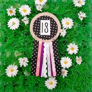 Op de afbeelding: Een zwarte en witte polka dot stof rosette met een jute centrum en een wit vierkant met het nummer 18 erop gedrukt. De rosette is omringd door witte madeliefjes en groen gras.