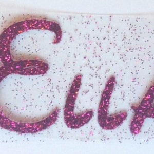 K&ouml;nnte beinhalten: Ein individueller Aufb&uuml;gler mit dem Namen Ella in einer geschwungenen Schreibschrift. Die Buchstaben bestehen aus einem leuchtend magentafarbenen Glitzermaterial, und die transparente Klebefolie ist mit feinen magentafarbenen Glitzerpartikeln &uuml;bers&auml;t.
