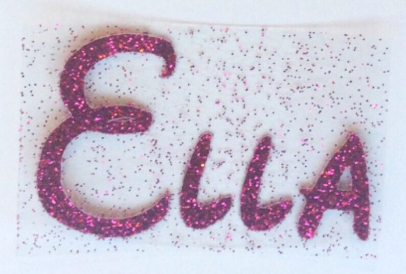 K&ouml;nnte beinhalten: Ein individueller Aufb&uuml;gler mit dem Namen Ella in einer geschwungenen Schreibschrift. Die Buchstaben bestehen aus einem leuchtend magentafarbenen Glitzermaterial, und die transparente Klebefolie ist mit feinen magentafarbenen Glitzerpartikeln &uuml;bers&auml;t.