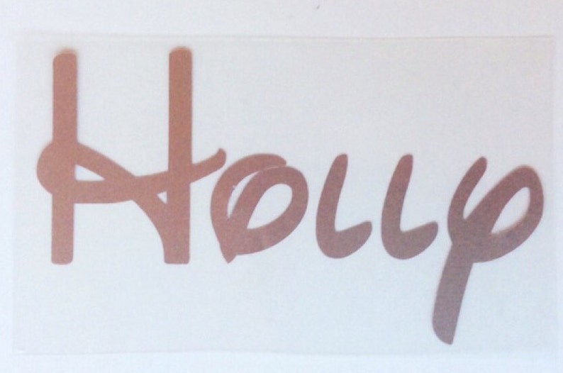 K&ouml;nnte beinhalten: Ein kupferfarbener Vinyl-Aufkleber mit dem Namen Holly in einer stilisierten Schreibschrift. Die Buchstaben sind zu einem einzigen St&uuml;ck verbunden und weisen ein metallisches Finish sowie glatte, abgerundete Kanten auf.
