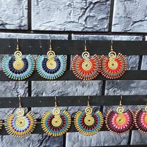 Puede incluir: Una colección de pendientes de macramé hechos a mano en varios colores, incluyendo azul, naranja, morado y rosa. Cada pendiente presenta un diseño de espiral dorada y flecos de cuentas. Los pendientes se exhiben en un soporte negro.