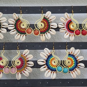 Könnte beinhalten: Eine Sammlung handgefertigter Ohrringe im Bohème-Stil. Jeder Ohrring hat ein spiralförmiges Design aus goldfarbenem Metall, einen zentralen Stein und ein Makramee-Muster, umgeben von Muscheln. Die Ohrringe sind in verschiedenen Farben erhältlich, darunter Gelb, Orange, Rot, Rosa und Türkis.