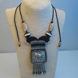 May include: A handcrafted necklace with a central rectangular pendant featuring an elephant design. The pendant is adorned with intricate floral patterns and dangling silver chains. The necklace has a black and gray macrame design with silver and wooden beads.