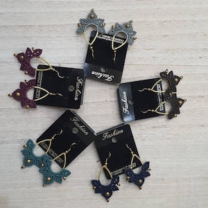 May include: A collection of handmade macrame earrings in various colors, including teal, navy, gray, and burgundy. Each pair features a unique geometric design with gold-colored metal accents and ear wires. The earrings are displayed on black cards labeled "Fashion".