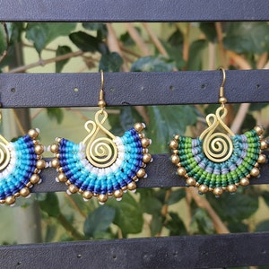 以下が含まれることがあります： 手作りのマクラメイヤリング4組。2組は青と白の色調で、2組は緑とグレーの色調です。各イヤリングには、金色のスパイラルと金色のビーズが付いています。