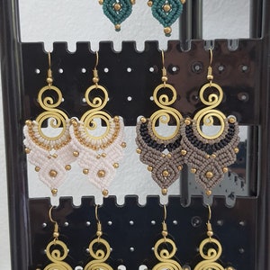 Puede incluir: Exhibición de pendientes de macramé hechos a mano en varios colores, incluyendo verde azulado, blanco, negro, rojo y beige. Cada par presenta una parte superior en espiral dorada y detalles de cuentas, mostrando un estilo bohemio.