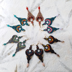 May include: A collection of handmade macrame earrings in various colors, including teal, brown, black, and gray. Each earring features intricate knotting, bead accents, and a unique teardrop shape. The earrings are displayed on a white surface.