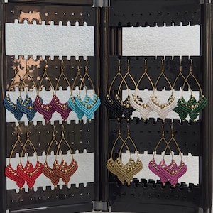 Puede incluir: Exhibición de pendientes de macramé hechos a mano en varios colores, incluyendo turquesa, burdeos, negro, blanco y verde. Cada pendiente presenta un alambre de latón en forma de lágrima y está adornado con pequeñas cuentas doradas. Los pendientes se exhiben en un expositor negro.