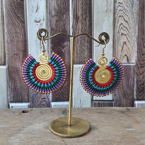 Puede incluir: Un par de pendientes de macramé coloridos con un diseño en espiral. Los pendientes presentan hilos rojos, azules, verdes y morados, acentuados con cuentas doradas y un centro en espiral dorado. Los pendientes se exhiben en un soporte dorado.