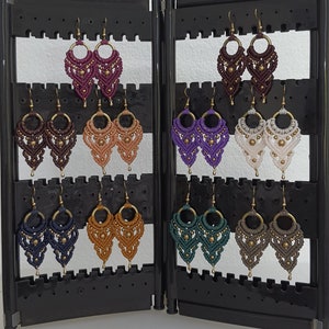 May include: A selection of handmade macrame earrings in shades of burgundy, brown, peach, purple, white, navy, gold, teal, and grey. Each pair features a circular top and detailed knotted patterns with gold-coloured accents.