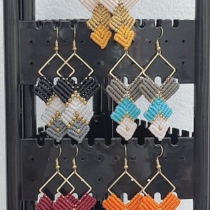May include: A selection of macrame earrings in a range of colours, including cream, gold, black, grey, teal, burgundy, and orange. Each pair has a geometric design with gold-coloured metal accents and ear wires. The earrings are displayed on a black earring stand.