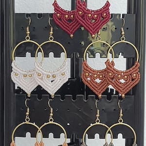 May include: A selection of handmade macrame hoop earrings in shades of burgundy, white, brown, peach, and grey. Each pair features a gold-coloured hoop and decorative macrame detailing with small gold beads. The earrings are displayed on a black earring holder.