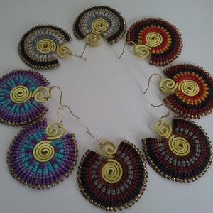 Puede incluir: Una colección de pendientes hechos a mano en varios colores, incluyendo morado, turquesa, rojo y gris. Cada pendiente presenta un diseño en forma de abanico con un acento de latón en espiral y bordes con cuentas. Los pendientes están dispuestos en un patrón circular.