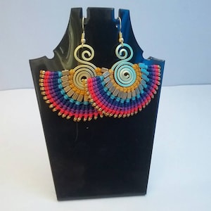 May include: A pair of vibrant macrame earrings. They have a fan shape with a brass spiral top and a fringe of red, purple, blue, and yellow threads. Small gold beads are at the bottom. The earrings are displayed on a black stand.