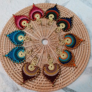 May include: A selection of handmade macrame earrings in shades of red, blue, and brown. Each earring has a leaf shape with a spiral centre and gold-coloured metal details. The earrings are presented on a woven mat.