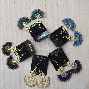 Puede incluir: Un conjunto de pendientes hechos a mano en varios colores, incluyendo verde azulado, azul, negro, crema y morado. Cada pendiente presenta un diseño en forma de abanico con intrincados detalles de macramé y acentos de alambre dorado. Los pendientes se exhiben en tarjetas negras.