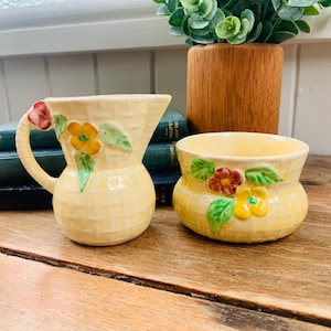 May include: A vintage yellow ceramic creamer and sugar bowl set. The pieces have a woven basket texture and are decorated with colourful flowers and green leaves. The creamer has a handle and a spout.