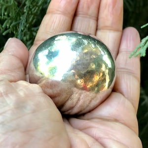 Könnte beinhalten: Eine kleine, silberfarbene, gehämmerten Metallkugel. Die Oberfläche reflektiert das Licht und erzeugt ein helles, glänzendes Aussehen. Die Kugel wird in einer offenen Hand gehalten, mit einem grünen, blättrigen Hintergrund.