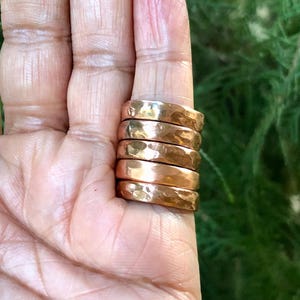 Op de afbeelding: Zes gehamerde koperen ringen gestapeld op een vinger. De ringen hebben een getextureerd oppervlak en een warme metallic kleur. De achtergrond is een vage groene blad. Elke ring heeft een diameter van ongeveer 2 cm.