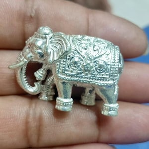 May include: A detailed silver elephant figurine with intricate carvings on its body and trunk. The elephant has a decorative blanket and is shown in a close-up shot. The figurine is held in a person's hand.