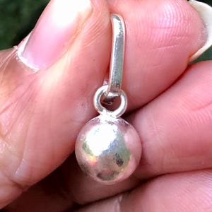 May include: A small, silver charm featuring a spherical bead and a loop for attachment. The charm is held in a hand, with a blurred green background.