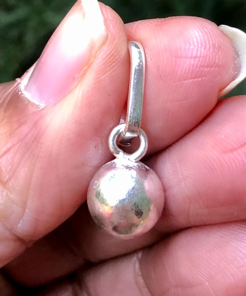 May include: A small, silver charm featuring a spherical bead and a loop for attachment. The charm is held in a hand, with a blurred green background.