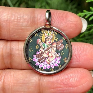 May include: A round silver pendant featuring a colorful illustration of a deity seated on a lotus flower. The artwork includes gold, green, pink, and brown hues against a dark green background. The pendant has a silver bail for attaching to a chain.