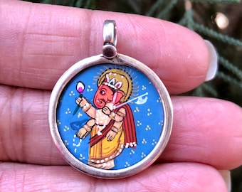May include: A silver pendant with a blue enamel background and a hand-painted image of the Hindu deity Ganesha. Ganesha is depicted in red and yellow with a pink lotus flower in his hand. The pendant is circular and has a silver bail.
