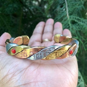 Puede incluir: Brazalete de puño de metal trenzado con tonos dorados, plateados y cobrizos. El brazalete presenta detalles rojos y símbolos grabados. El brazalete se sostiene en una mano sobre un fondo verde borroso.