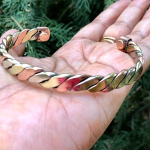 Op de afbeelding: Een gedraaide metalen armband met koper-, goud- en zilverkleurige tinten. De armband wordt in een hand gehouden, waarbij het ingewikkelde ontwerp en de kleuren worden benadrukt. De sluiting is zichtbaar, wat bijdraagt aan de functionaliteit.