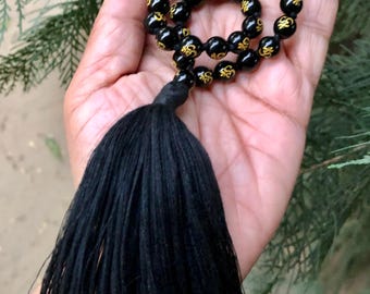 May include: Black beaded mala prayer beads with gold-colored Om symbols. The beads are held in a hand, with a long black tassel hanging from the bottom. The background is a blurred view of green foliage.