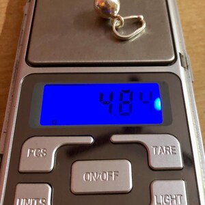 May include: A silver earring with a spherical design is placed on a digital scale. The scale reads 4.8 grams. The scale has buttons for power, units, and other functions. The background is a wooden surface.