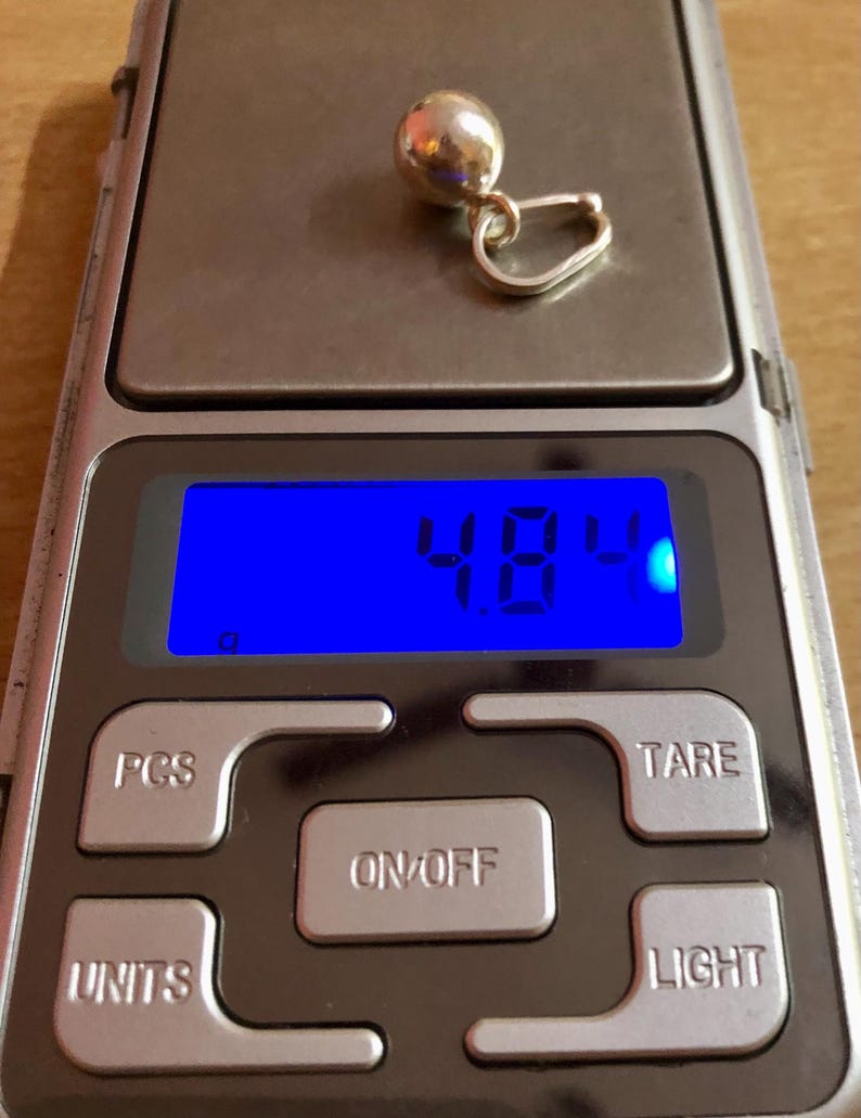 May include: A silver earring with a spherical design is placed on a digital scale. The scale reads 4.8 grams. The scale has buttons for power, units, and other functions. The background is a wooden surface.