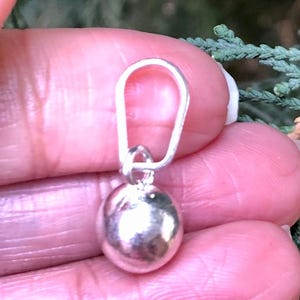 May include: A small, silver pendant with a rounded, pebble-like form. The pendant is connected to a silver loop, suitable for a necklace or bracelet. The jewellery is displayed against a blurred background.
