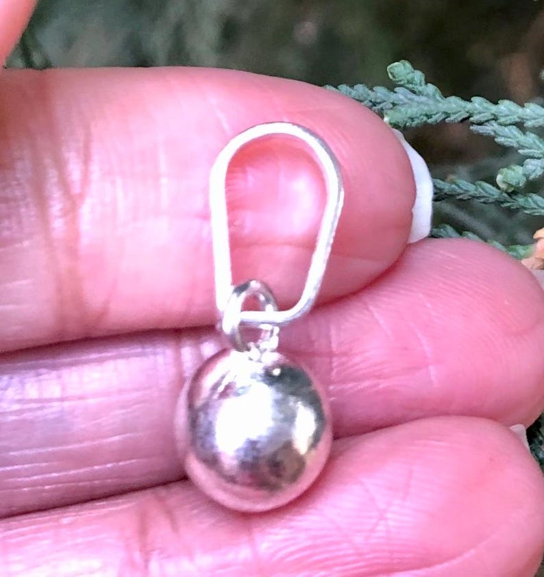 May include: A small, silver pendant with a rounded, pebble-like form. The pendant is connected to a silver loop, suitable for a necklace or bracelet. The jewellery is displayed against a blurred background.