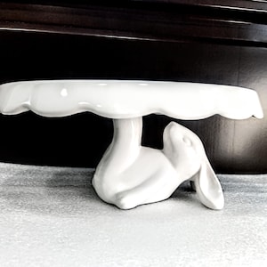 Puede incluir: Un soporte para tartas de cerámica blanca con un borde festoneado y una base de conejo. El soporte es perfecto para exhibir postres u otras golosinas.