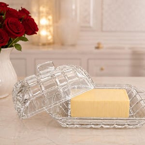 Puede incluir: Una mantequillera de cristal transparente con un patrón de rejilla cuadrada en la tapa y la base. La tapa está parcialmente levantada, revelando un bloque rectangular sólido de mantequilla amarilla sobre la base. La tapa tiene un asa pequeña.