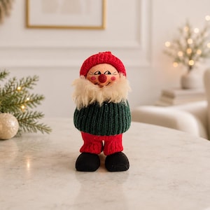 May include: A Christmas gnome doll with a red knitted hat and trousers, a green knitted sweater, and a fluffy white beard. The doll has a smiling face and black shoes. This festive decoration is ideal for adding a touch of whimsy to any home.