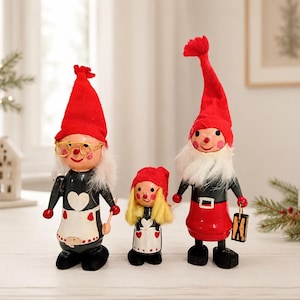 May include: Three decorative figurines featuring two tall figures and one smaller figure in the center. All wear pointed red hats and have white beards. The figures are dressed in black and red outfits with white aprons featuring heart patterns.