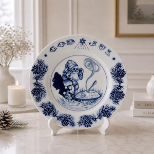 May include: A white ceramic decorative plate featuring a blue illustration of a gnome eating from a bowl. The rim is decorated with pine branches and cones, with the text "GOD JUL" and a star at the top. The plate rests on a white stand.
