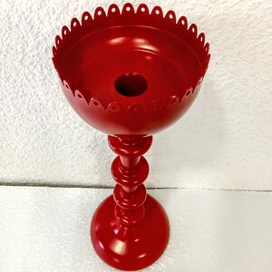 May include: A bright red metal candle holder featuring a tall, slender stem with multiple stacked circular rings and a wide, bowl-shaped top. The upper rim of the candle holder is finished with a decorative scalloped edge, providing a distinct silhouette.