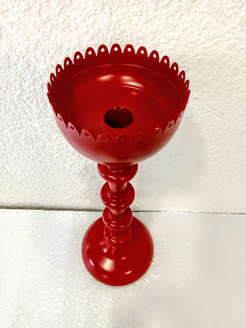 May include: A bright red metal candle holder featuring a tall, slender stem with multiple stacked circular rings and a wide, bowl-shaped top. The upper rim of the candle holder is finished with a decorative scalloped edge, providing a distinct silhouette.