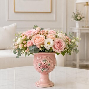 Op de afbeelding: Een roze keramische vaas gevuld met een boeket van roze en witte rozen, anjers en groen. De vaas heeft een voetstuk en een bloemenreliëf. De opstelling staat op een witte marmeren tafel.
