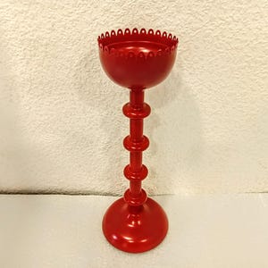 A red metal candle holder with a scalloped top and a tall, slender base. The base consists of stacked cylindrical sections, creating a visually interesting design. The overall style is simple yet elegant, suitable for various home decor settings.  The red finish is smooth and uniform, suggesting a high-quality paint job. This decorative piece would add a touch of modern style to any room.