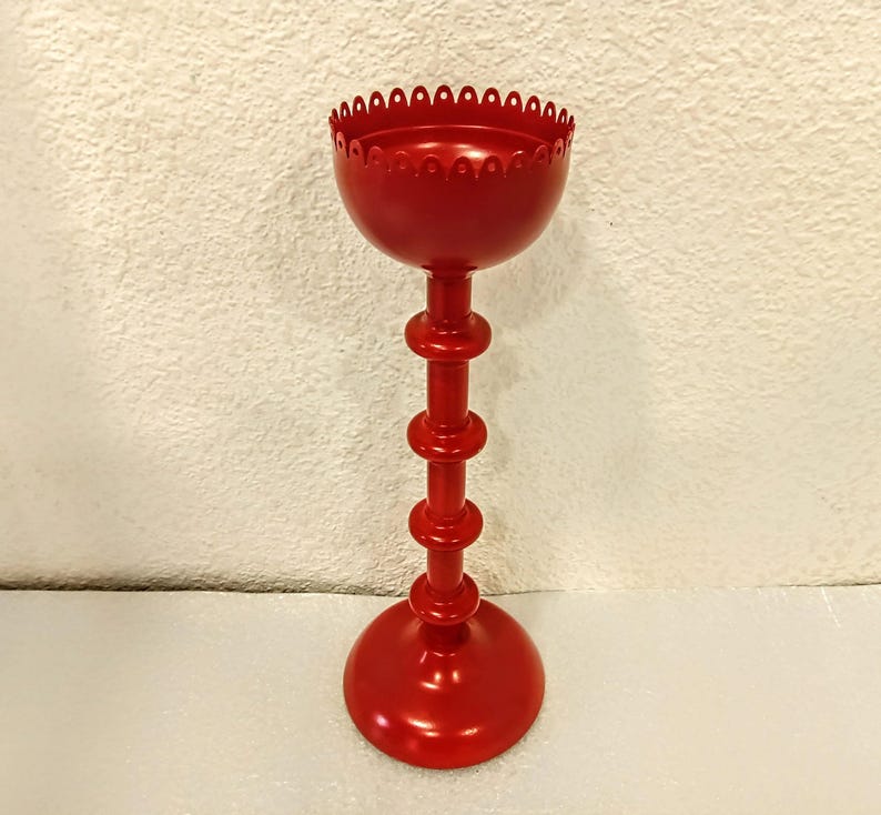 A red metal candle holder with a scalloped top and a tall, slender base. The base consists of stacked cylindrical sections, creating a visually interesting design. The overall style is simple yet elegant, suitable for various home decor settings.  The red finish is smooth and uniform, suggesting a high-quality paint job. This decorative piece would add a touch of modern style to any room.