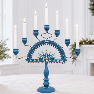 May include: A blue wooden candelabra featuring a central arched design with a starburst motif, painted with white and pink decorative patterns. It holds seven white taper candles in individual blue cups supported by curved metal arms.