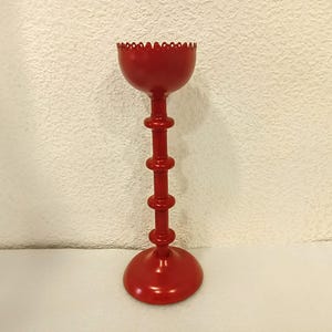 May include: A bright red metal candle holder featuring a tall, slender stem with three distinct horizontal ring accents. The top section is a bowl-shaped cup with a scalloped, decorative edge, and the base is a wide, circular, flat disc.